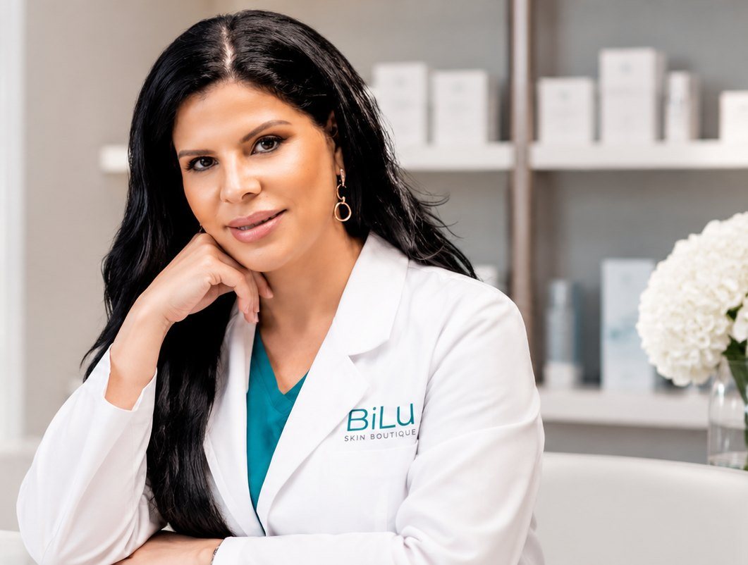 Luisa Barón, founder of BīLu Skin Health Boutique, in a white clinical coat, smiling with her chin resting on her hand